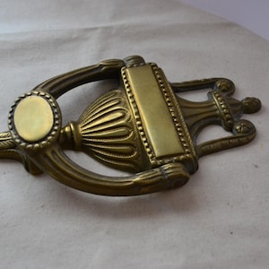 Puede incluir: Un llamador de puerta de latón con un diseño ornamentado. El llamador tiene una placa circular con un borde elevado y un cuerpo decorativo en forma de urna.