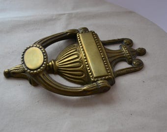 May include: A brass door knocker with an ornate design. The knocker features a circular plate with a raised rim and a decorative urn-shaped body.