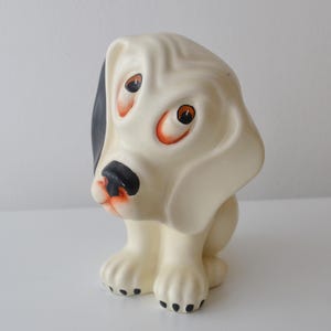 Könnte beinhalten: Eine cremefarbene Hundefigur mit langen, hängenden Ohren und einem traurigen Gesichtsausdruck. Der Hund hat einen schwarzen Fleck an einem Ohr, eine schwarze Nase und orange umrandete Augen. Die Pfoten sind mit kleinen schwarzen Punkten versehen.