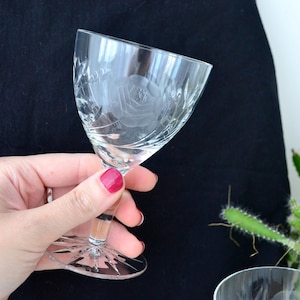 Peut inclure: Un verre clair gravé avec un motif floral de rose. Le verre a une base à pied avec un motif en étoile. Le verre est tenu sur un fond sombre, mettant en valeur les détails du motif gravé.