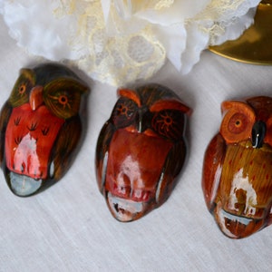 May include: Three wooden owl figurines painted in shades of brown, orange, and red. The owls have black eyes and beaks.