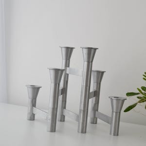 May include: A silver metal candle holder with six candle cups. The modern design features a geometric structure with vertical and horizontal bars. The candle holder is on a white surface.
