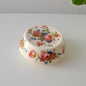 Puede incluir: Salero de cerámica color crema con un estampado floral de rosas rosas, amarillas y azules. El salero tiene forma redondeada con agujeros en la parte superior para dispensar la sal. Un artículo de cocina decorativo.