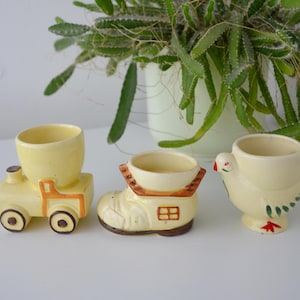 May include: Three vintage ceramic egg cups in the shape of a train, a boot, and a chicken. The egg cups are painted in shades of yellow, brown, and orange. The train has brown wheels, the boot has a small window, and the chicken has red and green accents.