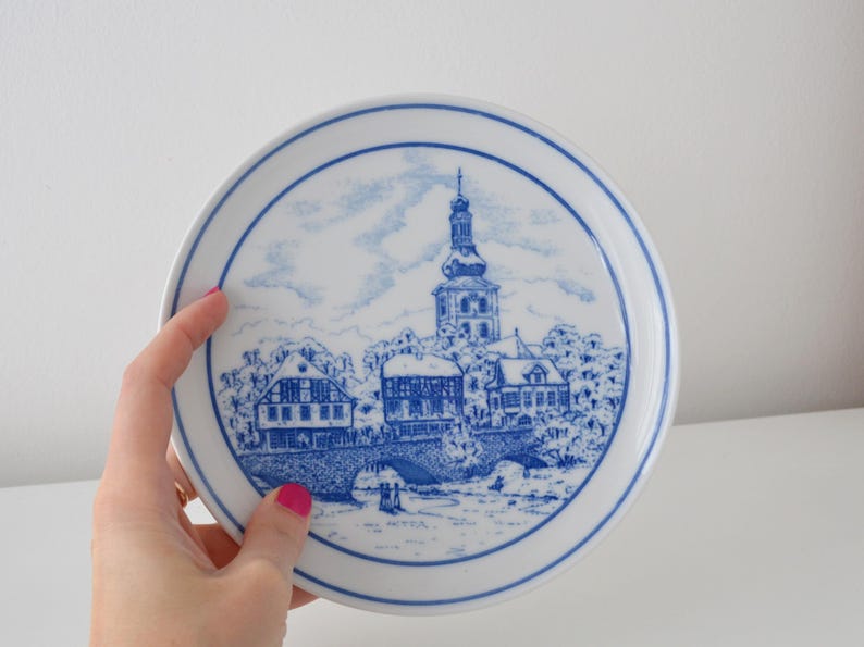May include: A white ceramic plate with a blue and white illustration of a European townscape. The plate features a detailed depiction of buildings, a bridge, and a church tower, all in a classic blue and white colour scheme. The plate has a blue border.