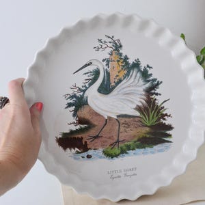 Può includere: Piatto da torta in ceramica bianca con bordo smerlato. Il piatto presenta un'illustrazione dettagliata di un airone bianco vicino a un ruscello, con il testo "Little Egret" sotto l'immagine. Lo sfondo include alberi e fogliame.