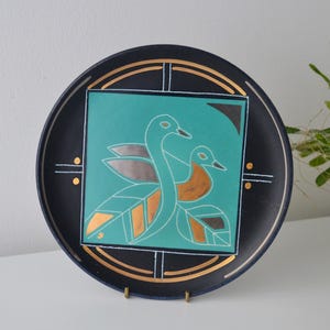 May include: Decorative black plate with a teal square featuring two stylized birds. The birds have orange and silver accents, with gold and white details. The plate has a gold rim and decorative gold accents.