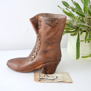 May include: A brown pottery boot with a lace-up design and small gold-colored studs. The boot has a small heel and is displayed with a tag that reads "HAND MADE POTTERY BOOTS". A green plant in a white pot is in the background.