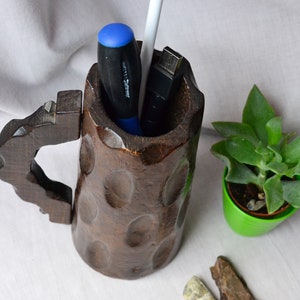 May include: A dark brown wooden mug with a carved handle, used as a desk organizer. It contains a blue screwdriver, a white pen, and a black USB drive.