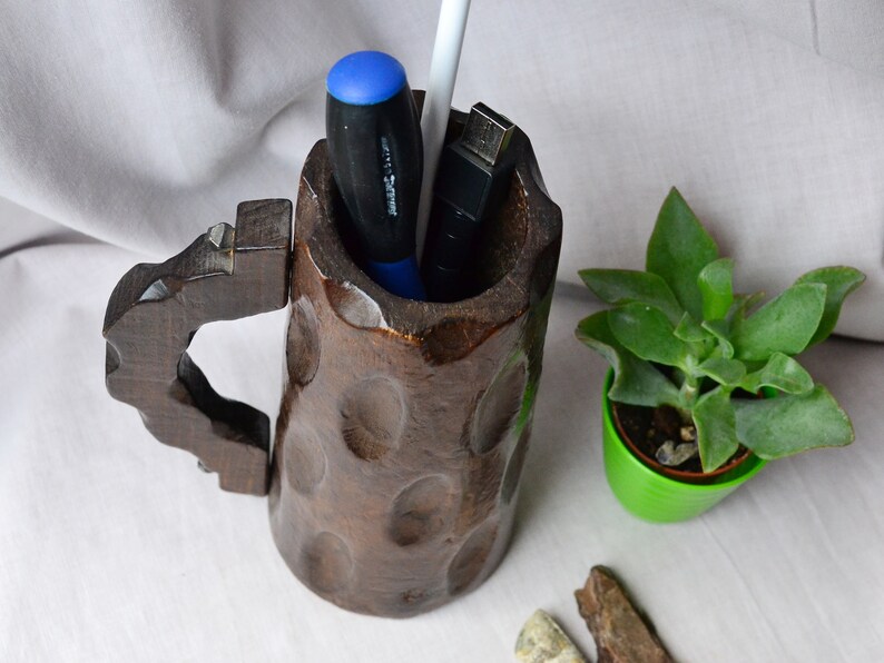 May include: A dark brown wooden mug with a carved handle, used as a desk organizer. It contains a blue screwdriver, a white pen, and a black USB drive.