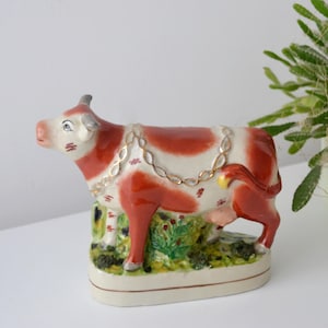 Op de afbeelding: Een keramieken koeienbeeldje met een rood en wit patroon, versierd met een goudkleurige ketting. De koe staat op een voet met groene en gele grasdetails. Het beeldje is ongeveer 15 cm lang.