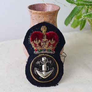 Puede incluir: Parche bordado azul marino con una corona de terciopelo rojo con detalles dorados sobre un emblema circular con un ancla plateada y una cuerda dorada. Insignia militar o náutica.