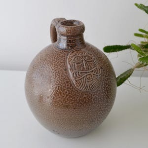 May include: A brown, round ceramic jug with a handle and a textured surface. The jug features a carved face design on the front. The jug is a decorative item.