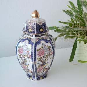 May include: A decorative ceramic jar with a lid, featuring a floral and bird design in pink, blue, and gold. The jar has a gold finial and is set against a white background.