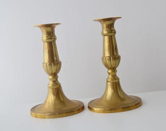 May include: A pair of polished brass candlesticks featuring a flared base with concentric ring detailing, a slender central column, and a fluted section above the base. The metallic gold-toned holders have a wide top rim for candles.