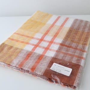 Könnte beinhalten: Eine gefaltete Tischdecke in einer transparenten Plastikverpackung. Die Tischdecke hat ein Karomuster in Gelb-, Orange-, Weiß- und Brauntönen. Das Etikett lautet "ONE TABLECLOTH".