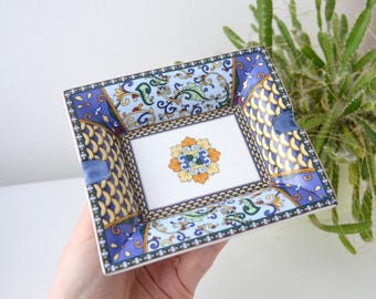 May include: Square ceramic ashtray with a decorative floral and scroll design. The ashtray features a white center with a yellow, orange, and blue floral motif. The border is blue with yellow and gold accents.