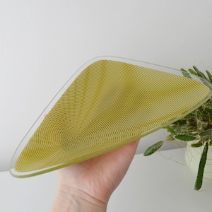 Könnte beinhalten: Eine transparente, dreieckige Glasschale mit gelb-grüner, strukturierter Innenseite. Die Schale hat ein einzigartiges Design mit konzentrischen Linien, die vom Zentrum ausgehen. Die Schale wird in einer Hand gehalten, mit weißem Hintergrund und einer Topfpflanze.