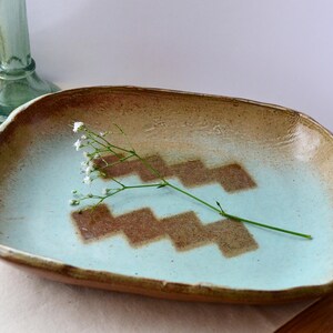 May include: A light blue ceramic serving dish with a brown geometric pattern. The dish has a slightly irregular shape and a rough texture. A sprig of white flowers is resting on the dish.