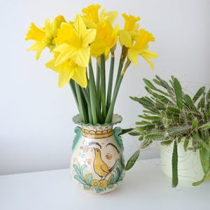 Könnte beinhalten: Ein Strauß gelber Narzissen in einer dekorativen Keramikvase. Die Vase ist cremefarben mit einem Vogelmuster und grünen und gelben Akzenten. Rechts befindet sich eine Topfpflanze.