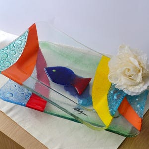 Puede incluir: Una colorida fuente de vidrio con diseño de pez. La fuente presenta un pez azul con aletas rojas y está decorada con detalles en naranja, amarillo y azul. Una flor blanca está adherida al lateral. La fuente mide unos 30 cm de largo.