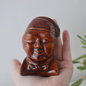 May include: A brown ceramic figurine of a smiling face wearing a hat and a bow tie. The vintage-style head is held in a hand, showcasing the details of the glazed finish. The figurine is a unique decorative item.