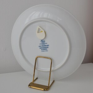 May include: A white ceramic plate with a blue and gold design, displayed on a gold-coloured metal stand. The plate has a small, white plastic hanger attached to the back. Text in blue is visible on the back of the plate.