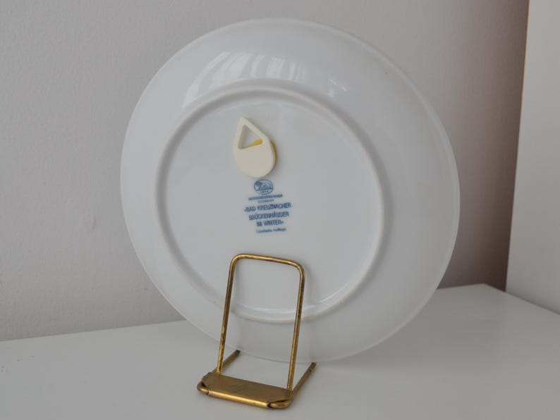 May include: A white ceramic plate with a blue and gold design, displayed on a gold-coloured metal stand. The plate has a small, white plastic hanger attached to the back. Text in blue is visible on the back of the plate.