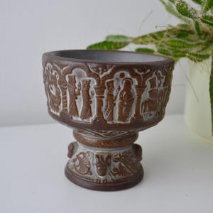 May include: A brown ceramic planter with a detailed relief design. The planter features a pedestal base and a wide rim, with a series of figures and architectural elements carved around the exterior. The interior is a darker brown color.