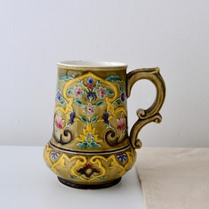 Op de afbeelding: Een antieke keramische mok met een decoratief bloemmotief in groen, geel, blauw en roze. De mok heeft een grote handgreep en een gedetailleerd, sierlijk patroon. De mok is ongeveer 15 cm hoog.