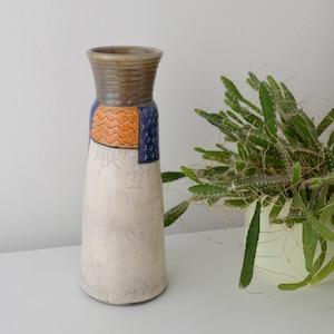 May include: A tall ceramic vase with a textured, off-white body. The top is brown with a ridged texture, and a section features orange and blue geometric designs. A green plant in a white pot is to the right.