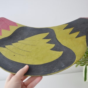 May include: A hand-painted, wooden chicken-shaped tray. The tray has a yellow body with black and pink accents. The head is black with a red wattle and yellow beak. A decorative piece for the kitchen or dining room.