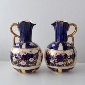 Könnte beinhalten: Zwei passende antike Keramikvasen. Jede Vase hat einen marineblauen Hals und Henkel mit goldenen Akzenten. Der Körper der Vasen ist weiß mit goldenen und floralen Designs. Die Vasen werden auf einer weißen Oberfläche präsentiert.