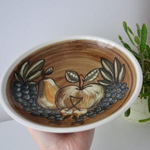 May include: Oval ceramic serving dish with a brown interior and white rim. The dish features a hand-painted design of fruit, including pears, apples, grapes, and cherries. The fruit is depicted in shades of brown, green, and white.