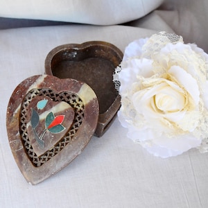 Puede incluir: Una caja de piedra tallada en forma de coraz&oacute;n con un dise&ntilde;o floral en rojo, azul y verde. La caja est&aacute; abierta y la tapa est&aacute; sentada a su lado. La caja est&aacute; sobre una superficie blanca con una flor de encaje blanco.
