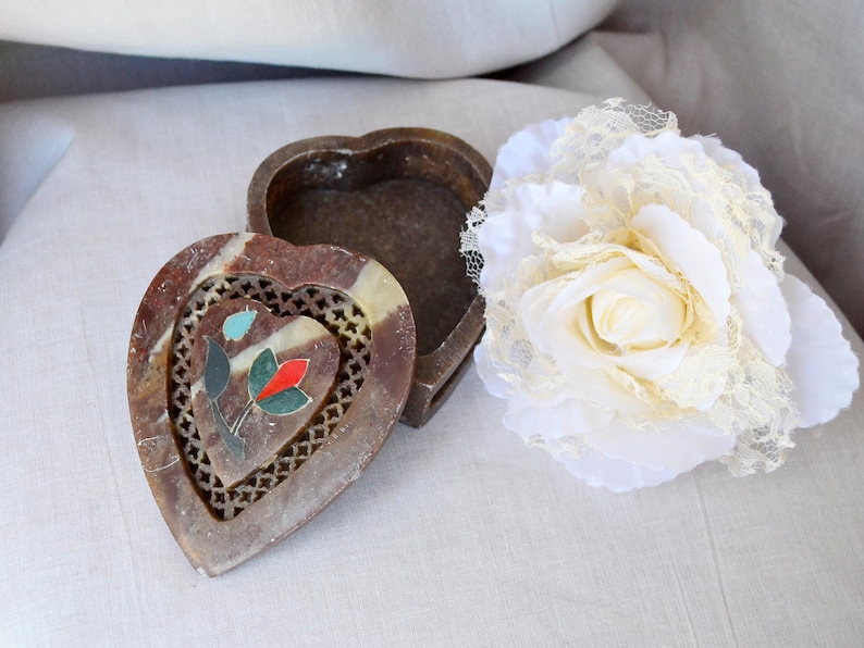 Puede incluir: Una caja de piedra tallada en forma de coraz&oacute;n con un dise&ntilde;o floral en rojo, azul y verde. La caja est&aacute; abierta y la tapa est&aacute; sentada a su lado. La caja est&aacute; sobre una superficie blanca con una flor de encaje blanco.
