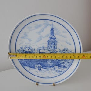 May include: White ceramic plate with a blue and white illustration of a European town scene. The plate is approximately 20 cm in diameter. The design features a church with a tall steeple and a bridge.