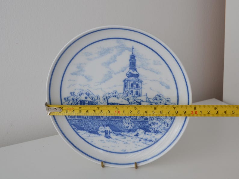May include: White ceramic plate with a blue and white illustration of a European town scene. The plate is approximately 20 cm in diameter. The design features a church with a tall steeple and a bridge.
