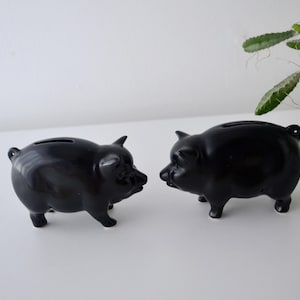 May include: Two glossy black ceramic piggy banks. Each bank has a slot on top for inserting coins and is shaped like a pig with four legs, a snout, and small ears. The banks are displayed on a white surface.