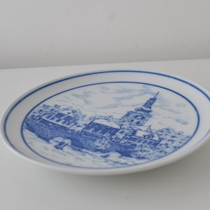 May include: White ceramic plate with a blue and white illustration of a European townscape. The plate features a detailed depiction of buildings, a bridge, and a tall church tower, all in a classic blue and white colour scheme.