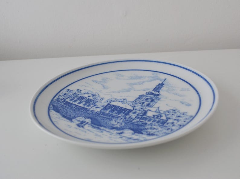 May include: White ceramic plate with a blue and white illustration of a European townscape. The plate features a detailed depiction of buildings, a bridge, and a tall church tower, all in a classic blue and white colour scheme.
