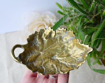 May include: A gold-toned brass leaf-shaped dish with a raised design. The dish has a handle on the side.