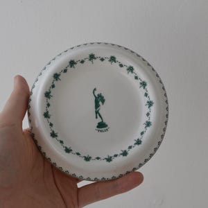 Może przedstawiać: Okrągły biały talerz ceramiczny z ciemnozielonym wzorem kwiatowym wzdłuż krawędzi. Na środku talerza znajduje się ciemnozielona sylwetka postaci z uniesioną ręką, umieszczona nad napisem "TOLLY" wykonanym zielonymi literami.
