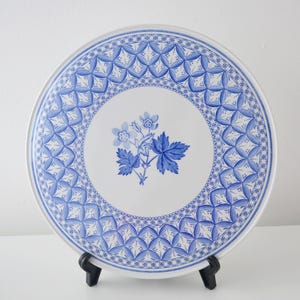 Può includere: Un piatto in ceramica bianca con un design floreale e geometrico blu. Il piatto presenta un motivo floreale centrale e un bordo a motivi. Il piatto è esposto su un supporto nero.