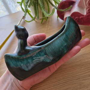 May include: A green and black ceramic canoe-shaped trinket dish with a cat figurine on the bow. The dish has a green glaze with a blue iridescent sheen. The text "Jameson 2-27" is written on the side of the dish.