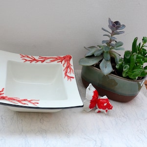 May include: A white square ceramic bowl with red coral designs and a black border. A small red and white goldfish figurine and a potted succulent plant are also present.