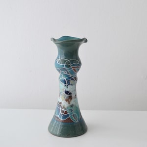 May include: A ceramic candlestick holder with a flared top and base, featuring a glossy teal glaze. The surface is decorated with a white, hand-painted linear pattern that creates a mosaic-like effect over patches of dark blue, brown, and white.