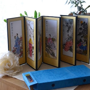 May include: A six-panel folding screen with traditional Asian artwork. Each panel features a different scene with figures in colorful robes, set against a gold background. A blue box is in the foreground.