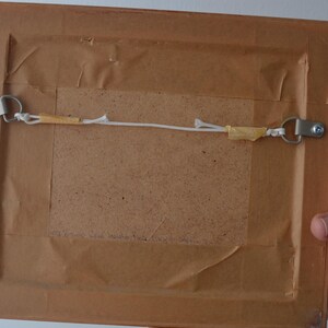 May include: The back of a rectangular picture frame featuring a brown paper backing, a central fiberboard panel, and two silver-toned metal D-ring hangers connected by a thin white string secured with small pieces of tan adhesive tape.