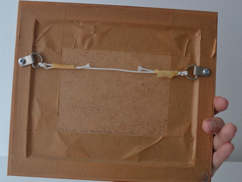 May include: The back of a rectangular picture frame featuring a brown paper backing, a central fiberboard panel, and two silver-toned metal D-ring hangers connected by a thin white string secured with small pieces of tan adhesive tape.